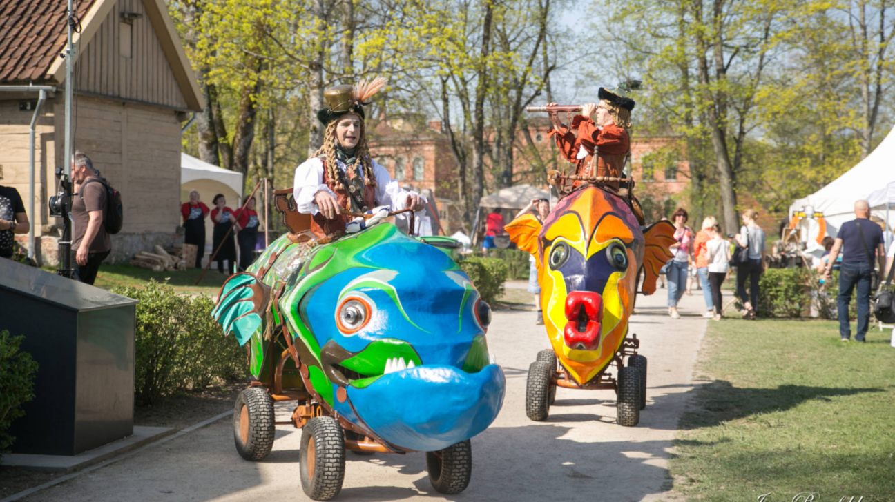 Divi teatrālos tērpos tērpti cilvēki brauc ar lieliem, krāsainiem zivju formas transportlīdzekļiem pa grants celiņu. Priekšplānā pa kreisi redzams priecīgs braucējs ar spalvainu cepuri un bizēm uz zili-zaļas zivs, bet labajā pusē – braucējs vēsturiskā tērpā, kurš caur teleskopu lūkojas uz priekšu no oranži-dzeltenas zivs. Fonā redzami cilvēki, zaļi koki un baltas teltis saulainā dienā. Ieva Benefelde PHOTOGRAPHY.
