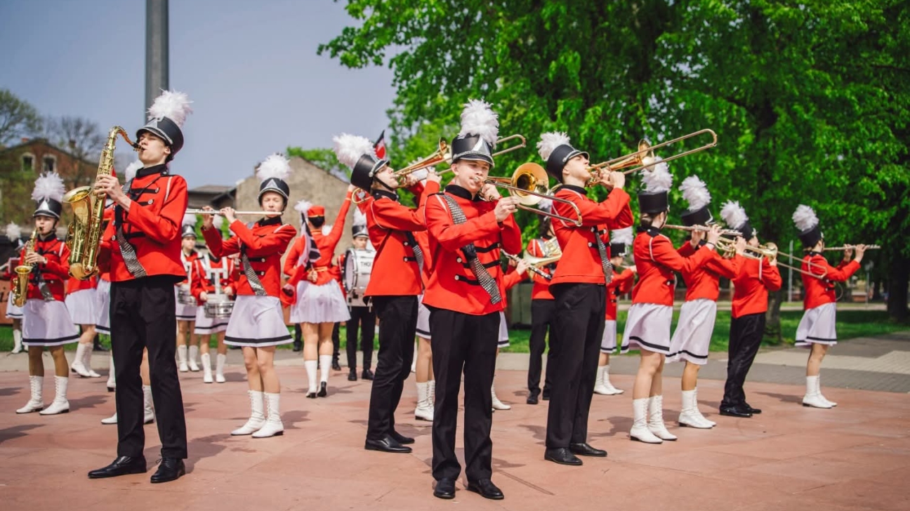 Maršējošs orķestris sarkanās un melnās uniformās ar baltām spalvu cepurēm spēlē pūšaminstrumentus, tostarp saksofonu, trombonus un flautas, ārā, ko ieskauj zaļi koki.