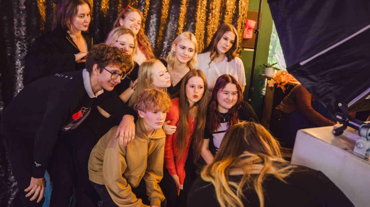 Desmit jaunieši pozē fotogrāfijai ar mirdzošu fonu un studijas apgaismojumu labajā pusē. Viņi stāv cieši kopā, daži smaida un skatās kameras virzienā.